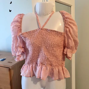 Rosa smockad topp öppna Axel  - Söt rosa topp med smockad design och volangdetalj i nederkant. Toppen har korta puffärmar och en halterneck-knytning. Perfekt för en somrig look!