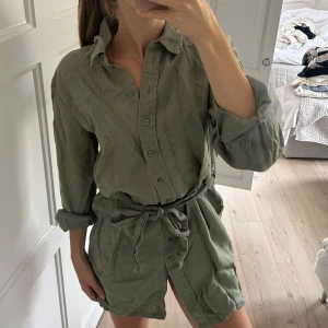 Grön skjortklänning med knytskärp från NA-KD - Snygg grön skjortklänning från NA-KD i linne med knytskärp i midjan. Klänningen har långa ärmar som kan vikas upp och knäppning framtill💕