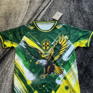  Brasilien fotbollströja  - Säljer en grön och gul fotbollströja med stort örnmotiv på framsidan och CBF Brasil-logga. Tröjan har korta ärmar, rund hals och detaljer i vitt och gult. Perfekt för dig som vill sticka ut på planen eller heja på Brasilien.