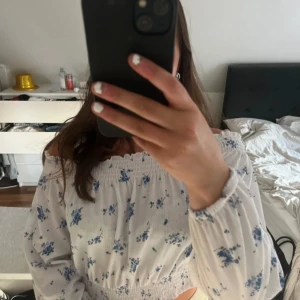 Vit offshoulder blus med blå blommor - Säljer en söt offshoulder blus i vitt med blått blommönster. Blusen har långa ärmar med resår vid ärmslut och midja, vilket ger en fin volangkant. Perfekt för vår och sommar!