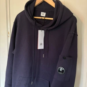  C.P. Company hoodie - Säljer en mörkblå hoodie från C.P. Company med den klassiska linsen på ärmen. Tröjan har huva, lång ärm och en ficka på ärmen. Perfekt för dig som gillar trendiga stilen och vill ha något stilrent men ändå unikt. Endast använd ett par gånger💯