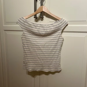 Vit offshoulder topp med svarta ränder - Säljer en vit ribbad offshoulder topp med tunna svarta horisontella ränder. Toppen har en bred, nedvikt halsringning och är figurnära i modellen. Perfekt till jeans eller kjol för en stilren look. I strl 36