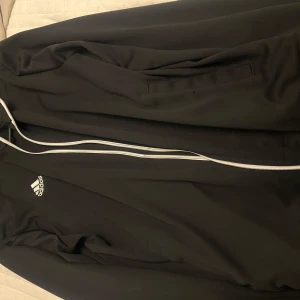Svart träningsjacka från Adidas - Säljer en svart träningsjacka från Adidas med vit dragkedja och logga på bröstet. Jackan har lång ärm och är perfekt för sport eller vardag. Klassisk och stilren design med fickor på sidorna.