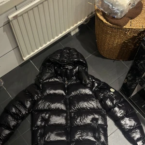 Moncler Maya - Säljer en snygg svart dunjacka från Moncler med glansig finish. Jackan har en huva och dragkedja framtill. Perfekt för kalla dagar.