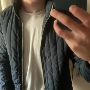 Mörkblå quiltad jacka från East West - Säljer en stilren mörkblå quiltad jacka från East West i storlek M. Jackan har dragkedja framtill och diskreta fickor på sidorna. Perfekt för dig som gillar en enkel och modern look. Ganska tunn, bör funka även på kallare sommar dagar