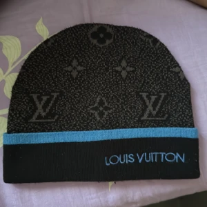 LV mössa - Snygg svart mössa från Louis Vuitton med klassiskt monogrammönster och blå kant. Broderad Louis Vuitton-logga framtill. Perfekt accessoar för att lyfta din stil.