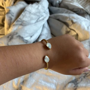 Guldfärgat armband med vita stenar - Säljer ett snyggt guldfärgat armband med två stora vita stenar i varje ände. Armbandet är ifrån Caroline Svedblom