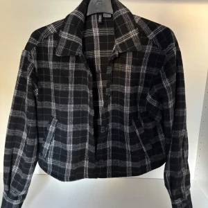 Svart och vit rutig croppad jacka från H&M Divided - Säljer en svart och vit rutig jacka från H&M Divided i croppad modell. Jackan har klassisk krage, knäppning framtill och långa ärmar. Perfekt för dig som gillar en trendig och avslappnad stil.