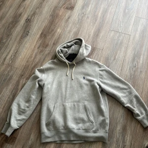 Grå hoodie från Polo Ralph Lauren - Säljer en klassisk grå hoodie från Polo Ralph Lauren med broderad logga på bröstet. Tröjan har huva med dragsko och en stor magficka framtill. Perfekt för en avslappnad stil.