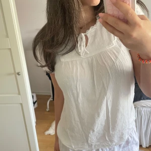 Vit blus med knyt i halsen - Säljer en söt vit blus med knyt vid halsen och lätt rynkade detaljer. Blusen har en luftig, avslappnad passform. Perfekt för varma dagar eller att styla lager på lager.