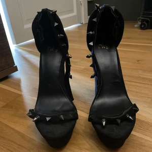 Svarta pumps med nitar från Nly - Säljer ett par svarta pumps från Nly med höga klackar och coola nitar längs remmarna och framtill. Skorna har en kraftig plattform och justerbara spännen vid ankeln. Perfekta för dig som vill sticka ut!