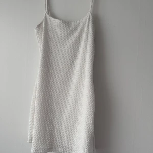 Vit kort klänning med struktur - Säljer en söt vit kortklänning med smala axelband och strukturerat tyg. Klänningen har en enkel och stilren design som passar perfekt till varma dagar. Lätt och luftig modell som ger en fräsch look.
