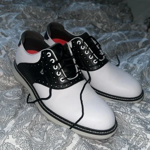 Svarta och vita golfskor från FootJoy - Säljer ett par klassiska golfskor från FootJoy i svart och vitt med snörning och röda detaljer på sulan. Skorna har rund tå och platt sula, perfekt för golfbanan. Använda 1 gång ute på banan så inprincip nya