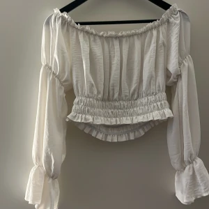 Vit offshoulder blus med volang - Säljer en söt vit offshoulder blus med långa puffärmar och smockad midja. Blusen har volangdetaljer vid halsringning, midja och ärmslut. Skriv för fler bilder!