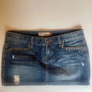 Lågmidjad jeanskjol med nitar - Lågmidjad cool mini jeanskjol med slitna detaljer och nitar längs fickorna framtill. Klassisk blå denim med råa kanter och femficksmodell. Perfekt för dig som gillar en edgy stil. Midjemått 41cm längd :30 cm