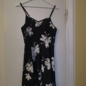 Svart blommig långklänning med smala axelband - Svart långklänning med stora vita och ljusblå blommor. Klänningen har smala axelband och v-ringad halsringning, perfekt för varma dagar. Lätt och luftig modell som ger en avslappnad men stilren look.