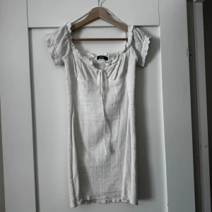 Vit kortklänning med puffärm från Bik Bok - Supersöt vit kortklänning från Bik Bok med puffärmar och smockad struktur. Klänningen har knytning vid bysten och volangdetaljer vid ärmslut och halsringning. Perfekt för sommarens alla tillfällen!