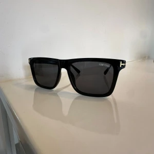 Svarta solglasögon från Tom Ford - Stilrena svarta solglasögon från Tom Ford med rektangulära bågar och mörka glas. Snygga gulddetaljer vid skalmarna och diskret logga på linsen. Tillverkade i Italien och passar perfekt för en trendig look.