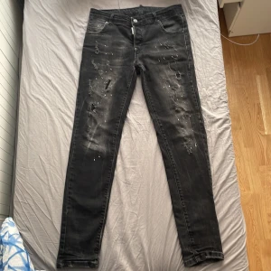 Svarta jeans från Dsquared2 med slitningar - Säljer ett par svarta jeans från Dsquared2 med slitna detaljer och färgstänk framtill. Jeansen har klassisk femficksdesign, knappgylf och smal passform. Perfekta för dig som vill ha en edgy och trendig look. Vet inte om den är äkta. Den är 97 cm lång.