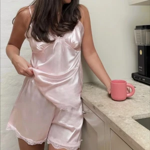 Mörkblå satinlinne och shorts-set med spets - Säljer ett mörkblått set i satin med linne och matchande shorts. Båda plaggen har tunna axelband och spetsdetaljer längs kanterna samt en liten rosett framtill på linnet. Perfekt för en lyxig känsla hemma.