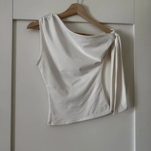 Vit oneshoulder topp med knyt - Snygg vit oneshoulder topp med knyt på ena axeln. Topp i mjukt material med figurnära passform, perfekt för att lyfta din outfit med en trendig touch.