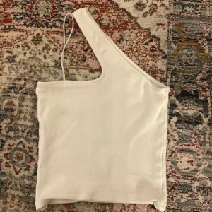 Vit oneshoulder ribbad topp - Säljer en vit ribbad oneshoulder topp med smal axelrem. Perfekt för dig som vill ha en stilren och trendig look. Toppens material är mjukt och stretchigt.