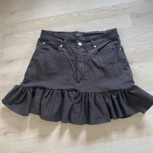 Svart volangkjol från Lagoon - Säljer en svart kjol från Lagoon med volangkant nertill. Kjolen har klassisk jeanslook med fickor fram och bak samt knapp och dragkedja i midjan. Perfekt för dig som gillar en cool och feminin stil.