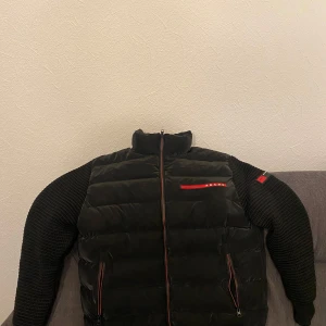 Svart pufferjacka från Prada - Säljer en svart pufferjacka från Prada med stickade ärmar och röda detaljer. Jackan har dragkedja framtill och två fickor med dragkedja. Klassisk Prada-logga på bröstet och ärmen. Storleken passar både M och L