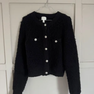 Svart cardigan jacka från H&M - Säljer en svart cardigan jacka från H&M med dekorativa silverfärgade knappar framtill och på fickorna. Jackan har lång ärm, rund halsringning och en fluffig, texturerad yta som ger en trendig look.