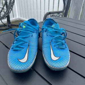 Blåa fotbollsskor Nike Phantom - Säljer ett par blåa Nike Phantom fotbollsskor med vita och svarta detaljer samt gröna dobbar. Skorna är bäst för gräs men funkar även på konstgräs. De är mottligt använda och jag är öppen för pris förhandling. 