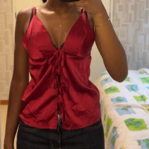 Röd omlottopp med knyt - Säljer en röd omlottopp, toppen har smala axelband, djup v-ringning och knyt framtill. Materialet är satin, ALDRIG ANVÄND, säljer då jag ångra mitt köp!