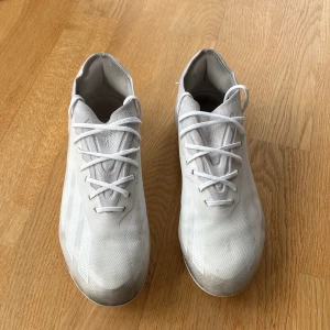 Vita fotbollsskor från Adidas (crazyfast) - Säljer ett par vita fotbollsskor från Adidas med låg profil och mönstrad ovandel. Skorna har rund tå och fasta dobbar för spel på gräs. Snygg minimalistisk design utan snörning och med diskreta Adidas-ränder på sidan.