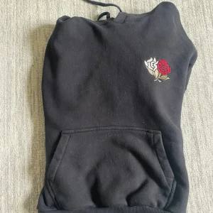 Svart hoodie med broderad ros från Les Deux - Svart hoodie från Les Deux med broderad röd och vit ros på bröstet. Tröjan har huva och en stor magficka framtill. Perfekt för en avslappnad och stilren look.