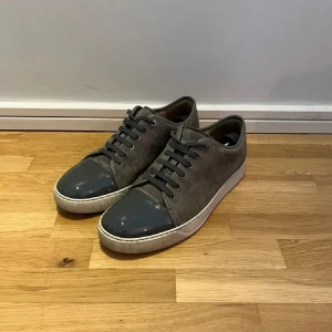Lanvins - Säljer ett par grå sneakers från Lanvin med snygg lackad tå i mörkgrått och ovandel i mocka. Skorna har snörning och vit platt sula. Perfekta för dig som gillar stilrena och exklusiva sneakers. Skorna har även ett litet hål i sulan, men inget som syns.