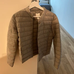 Beige pufferjacka från H&M - Säljer en beige pufferjacka från H&M i storlek 36. Jackan är lätt vadderad med rund halsringning och knäppning framtill. Perfekt för kyliga dagar och enkel att matcha med olika outfits.