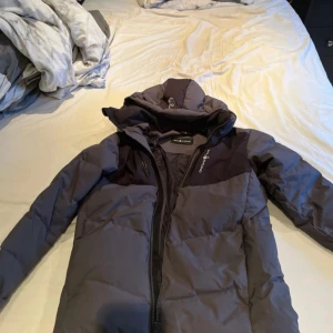 Grå dunjacka från Sail Racing - Säljer en grå dunjacka från Sail Racing med svart detalj över axlarna och logga på bröstet. Jackan har huva, dragkedja och är vadderad för extra värme. Perfekt för kyliga dagar.