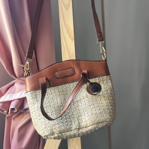 Beige och brun axelväska i strå och skinn - Säljer en snygg axelväska i beige stråmaterial med bruna skinn-detaljer. Väskan har både handtag och justerbar axelrem samt dragkedja upptill. Perfekt för dig som vill ha en stilren och somrig accessoar.
