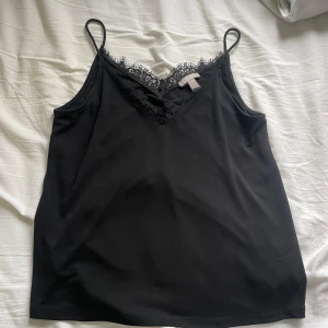 Svart linnetopp med spetsdetaljer - Säljer en svart linnetopp med tunna axelband och v-ringning dekorerad med spets. Toppen har en elegant och enkel design, perfekt att styla till olika outfits. Materialet känns mjukt och följsamt.