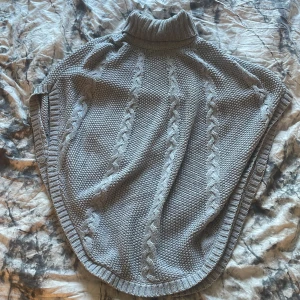 Grå stickad poncho med polokrage - Säljer en grå stickad poncho med polokrage och kabelstickat mönster. Perfekt att slänga över en tröja för extra värme. Mjuk och skön med breda kanter och knappdetaljer på sidorna.