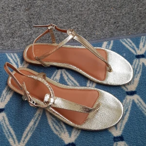 Guldiga sandaler från H&M - Säljer ett par snygga guldiga sandaler från H&M med smala remmar och spänne vid ankeln. Perfekta för sommaren och ger en glittrig touch till din outfit. Platt sula och öppen tå för maximal komfort. Skor är helt nya. 