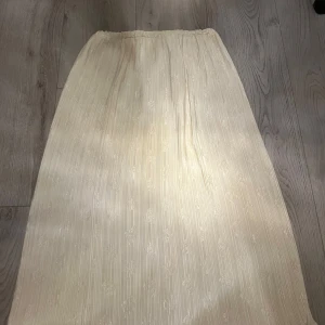 Plisserad beige långkjol knappast använd! - Säljer en plisserad beige långkjol med subtilt blommönster. Kjolen har resår i midjan och ett glansigt material som ger en elegant känsla. Perfekt för dig som gillar en stilren och feminin look.