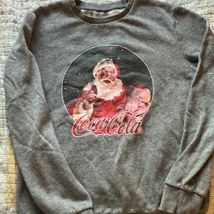 Grå sweatshirt med Coca-Cola jultomten - Grå sweatshirt med tryck av Coca-Cola jultomten på framsidan.  Bra för vintern och julen för dig som gillar klassiska motiv. Köpt på Göksäter.