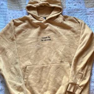 Orange/Gul hoodie från Black Squad. Tryck - färg prickar. - Köpte på en secondhand, Orange/gul oversized hoodie från Black Squad med svart text 'raised by the streets.' . Hoodien har tryckt färgstänk prickat mönster lite här och där över hela tröjan. Mjuk nopprig insida och ribbade muddar vid ärmslut och nederkant.