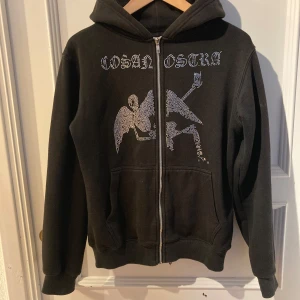 Svart hoodie med strasstryck från Cosa Nostra Stockholm - Svart hoodie med huva och dragkedja från Cosa Nostra Stockholm. Framsidan har ett stort strasstryck med text och motiv av en ängel och en figur som gör peace-tecken. Två fickor framtill och ribbade muddar.