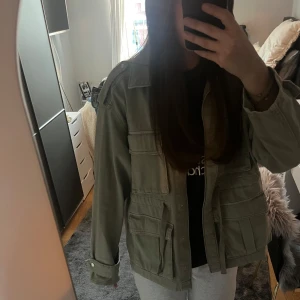 Grön jacka från H&M - Säljer en grön jacka från H&M Divided med flera stora fickor framtill och knappar. Jackan har klassisk krage och långa ärmar med manschett. Perfekt för lager-på-lager och enkel att styla till olika outfits.