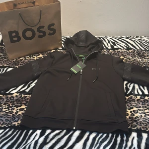 Svart kofta  från BOSS - Svart kofta från BOSS med dragkedja framtill och huva. Jackan har diskreta BOSS-loggor på ärmarna och bröstet samt två fickor fram. Snygga detaljer med mönstrade band längs ärmarna och huvan. Perfekt för en stilren och sportig look.