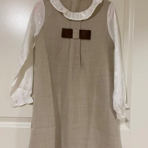 Beige klänning med vit blus och brun rosett - Söt beige klänning med vit blus under, volangkrage och långärmade ärmar med diskret blommönster. Framtill sitter en brun rosett som detalj. Perfekt för dig som gillar en klassisk och gullig stil.