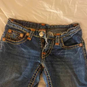 True religon jeans low waist bootcut - Säljer ett par jätteunika snygga mörkblå jeans från True Religion med tydliga orange kontrastsömmar och unika fickdetaljer. Dragkedjan på gylfen är sönder och utklippta där nere men annars skit snygga 