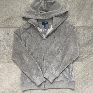 Helt ny Grå hoodie från Polo Ralph Lauren - Snygg grå hoodie från Polo Ralph Lauren i ribbad manchester med dragkedja framtill och huva. Helt ny! Klassisk broderad logga på bröstet. Perfekt för en avslappnad och stilren look. Storlek L 14-16 i denna modell motsvarar XS/S!👍