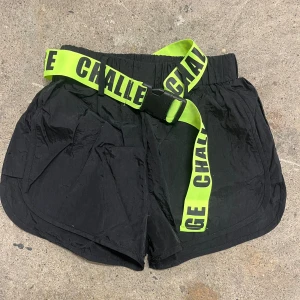 Svarta shorts med neongrönt bälte - Svarta syntetshorts från SHEIN med elastisk midja och ett neongrönt bälte med texten 'CHALLENGE'. Shortsen har sportig look och är perfekta för en trendig stil.